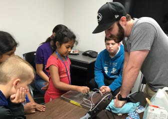 An child demonstrates weld test equipment as an instructor and other children look on.